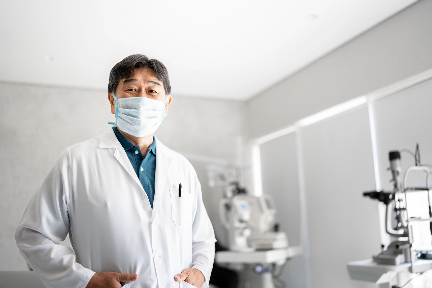 Eye care professional in lab coat and mask, standing in examination room