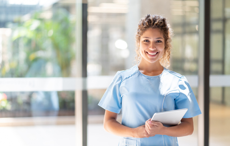 Woman in scrubs, in front of window and holding a tablet