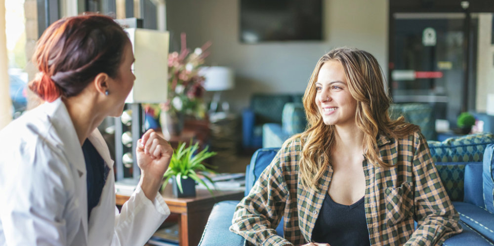 A healthcare professional in a white coat engages in conversation with a smiling woman in a plaid shirt, sitting in a cozy, well-lit waiting area with green plants and comfortable furniture.