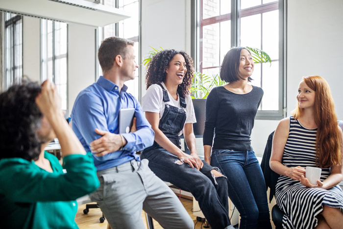 A diverse group of five people are seated in a circle, facing each other and laughing. They appear to be in a casual meeting or workshop setting.