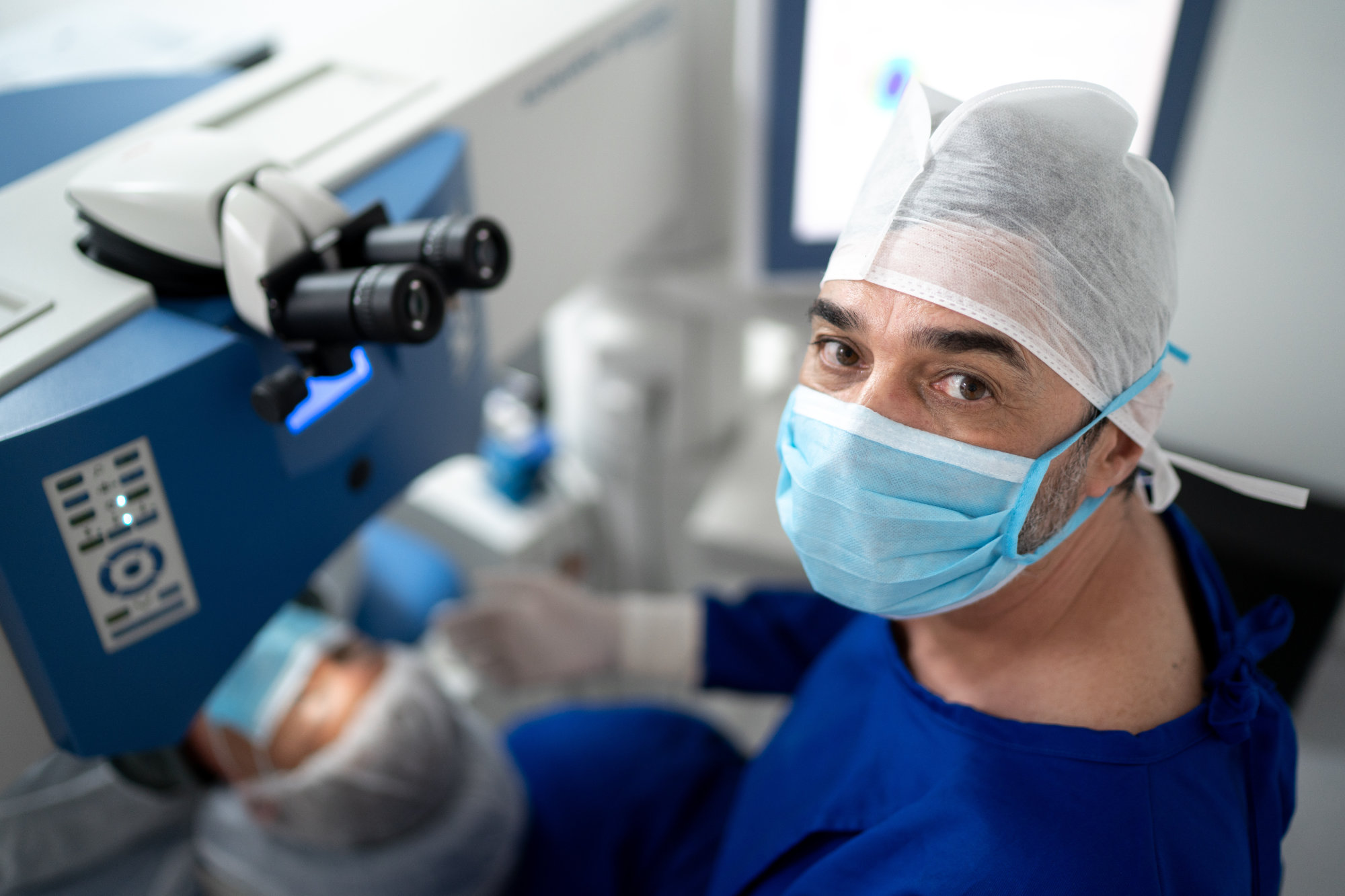 A close-up of an eye surgeon wearing a surgical cap and mask, looking directly at the camera with an eye scope in the background.
