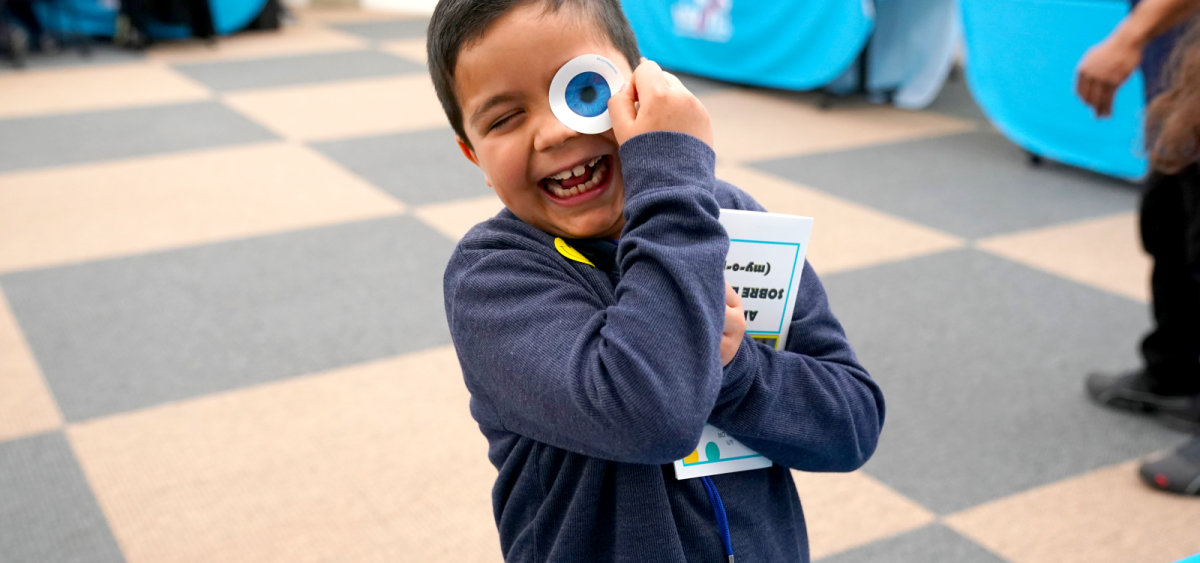 Smiling child holding a large googly eye toy up to their face while clutching a book outdoors.