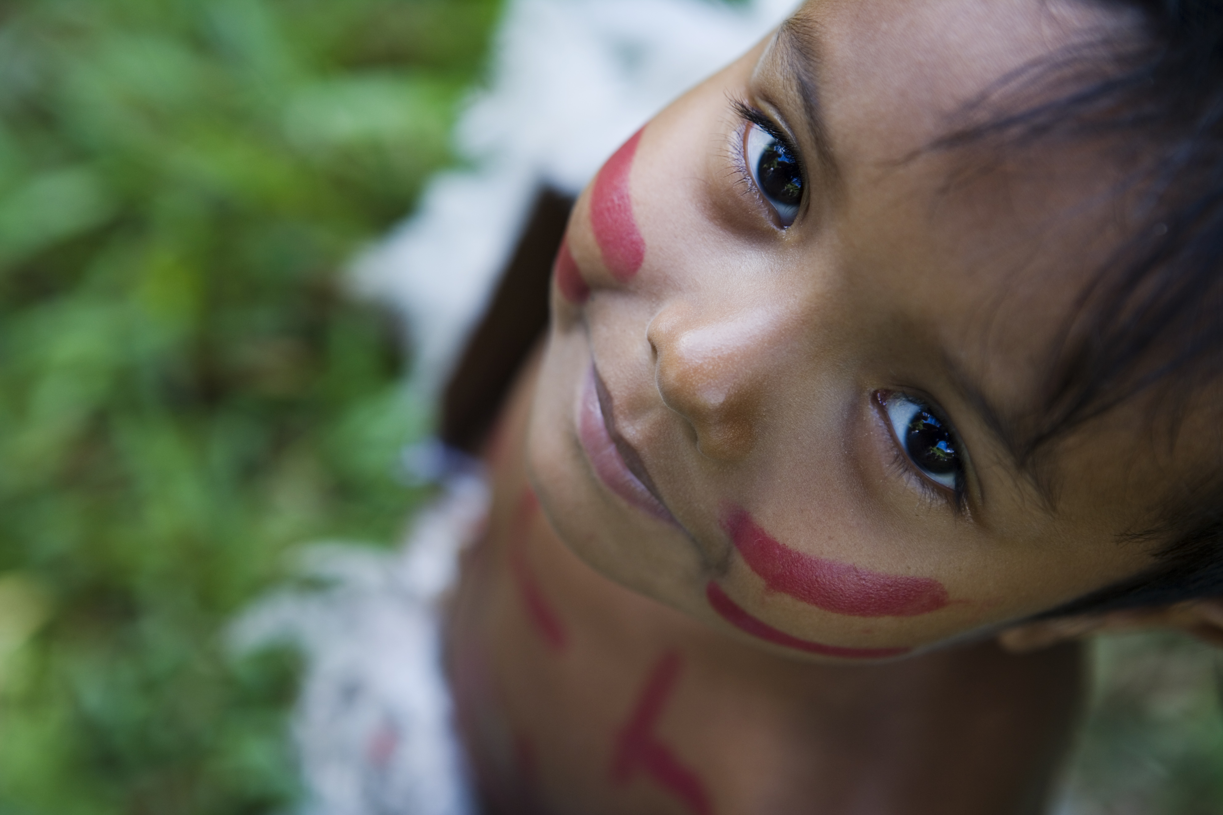 A child's face with red painted stripes on the cheeks is looking upward.
