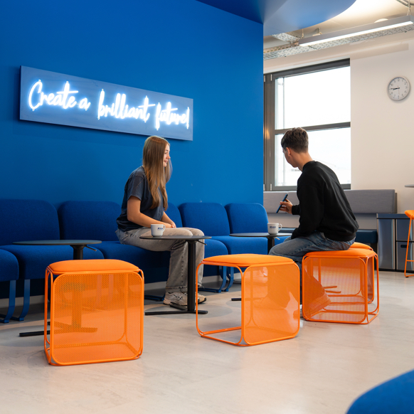 Modernes, helles Büro-Lounge-Bereich mit blauen Sitzmöbeln, orangefarbenen Hockern, in dem eine Trainerin und ein Auszubildender arbeiten, sowie ein leuchtendes Motivationsschild.