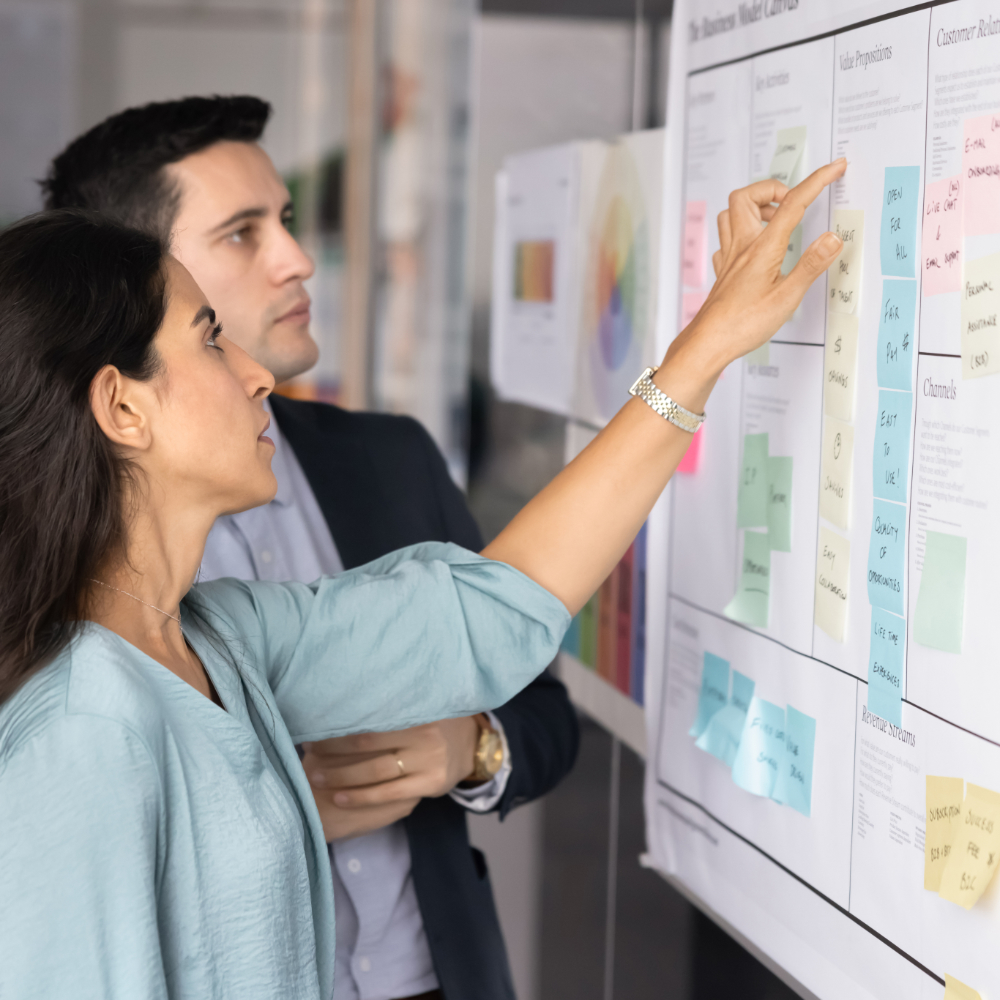 Zwei Kollegen überprüfen ein Strategieboard, das mit Haftnotizen bedeckt ist, und führen wahrscheinlich ein Brainstorming durch oder analysieren einen Plan.