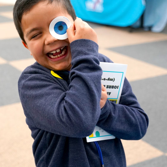 Smiling child holding a large googly eye toy up to their face while clutching a book outdoors.