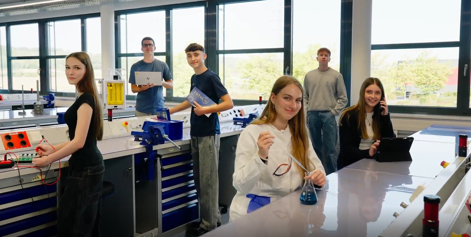 Eine Gruppe von sechs Schülern steht in einem hellen Wissenschaftslabor und hält verschiedene wissenschaftliche Werkzeuge. Sie wirken engagiert und selbstbewusst und vermitteln Teamarbeit und Lernbereitschaft.