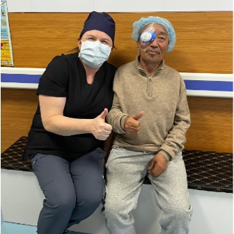 Patient with bandaged eye and medical professional in surgical mask sit together on bench and give a thumbs-up to the camera