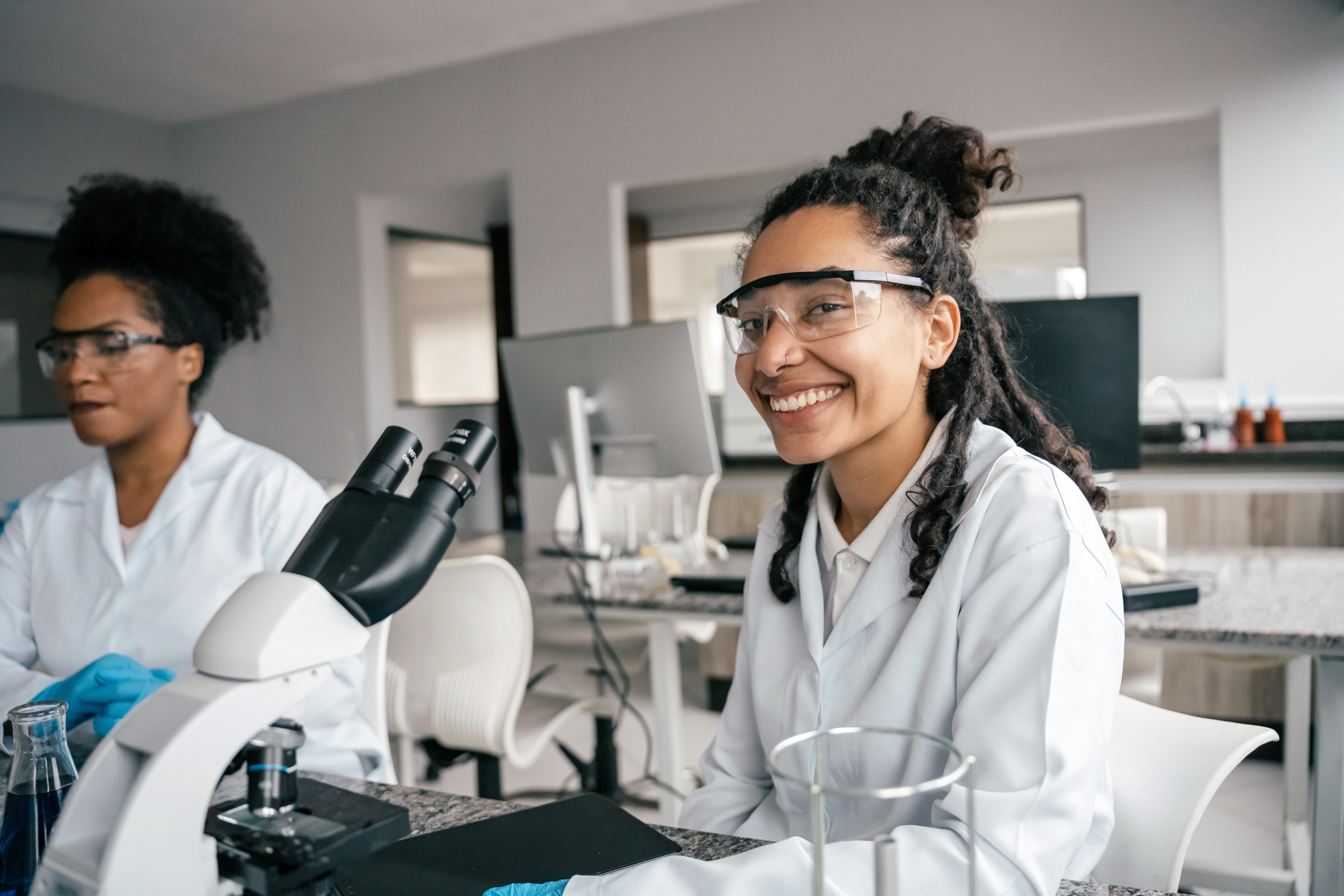 Two female scientists wearing lab coats,  safety glasses and gloves sitting at a table. one is sitting in front of a microscope and smiling at the camera.