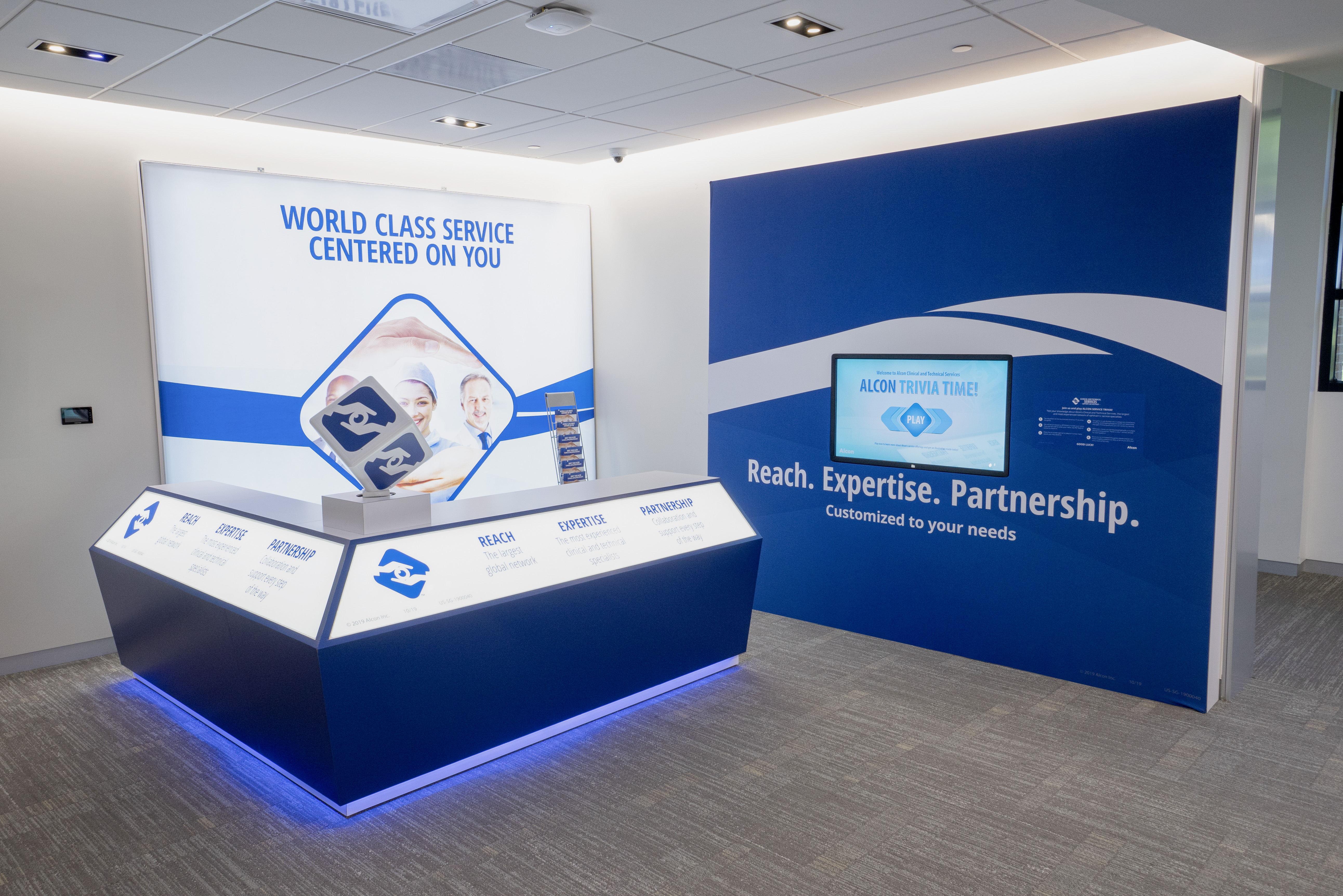 A blue and white branded display in a corporate office. The display features a digital screen with the text "Alcon Trivia Time!". The wall behind it reads "Reach. Expertise. Partnership. Customized to your needs."