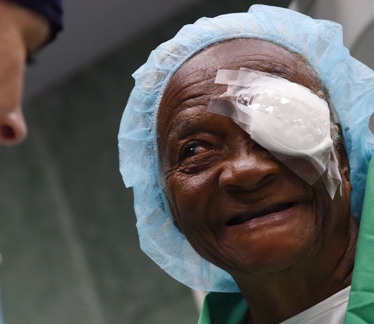 Primer plano de una persona mayor que lleva una cofia quirúrgica azul claro y un parche en un ojo. La persona sonríe suavemente. En el fondo, se ve parcialmente a otra persona desenfocada que lleva mascarilla quirúrgica y ropa verde de quirófano.