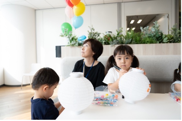 明るく広々とした空間で、大人が見守る中、子供が白いボールを持っています。背景には風船が見えます。