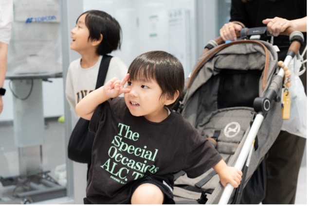 「The Special Occasions」とプリントされた黒いシャツを着た幼い男の子が、デジタルスクリーンを見上げています。背景にはベビーカーが見えます。