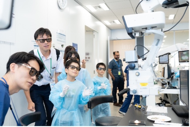 明るい医療研究室で、青い防護服を着た子供たちと大人がロボットアームを見学しています。