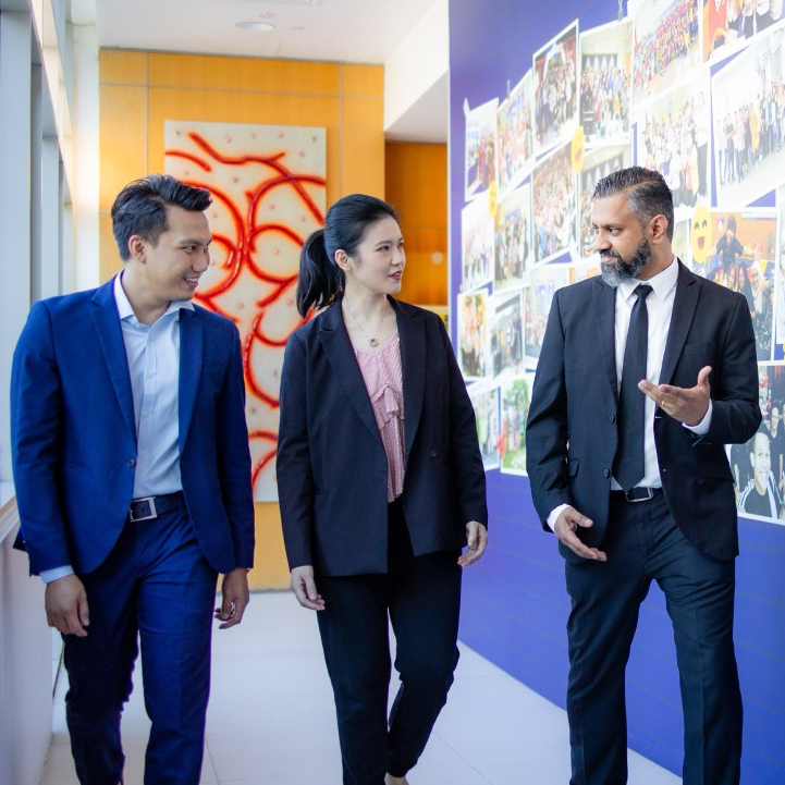 Three business professionals walking down a modern office hallway, engaged in conversation with a photo wall in the background.