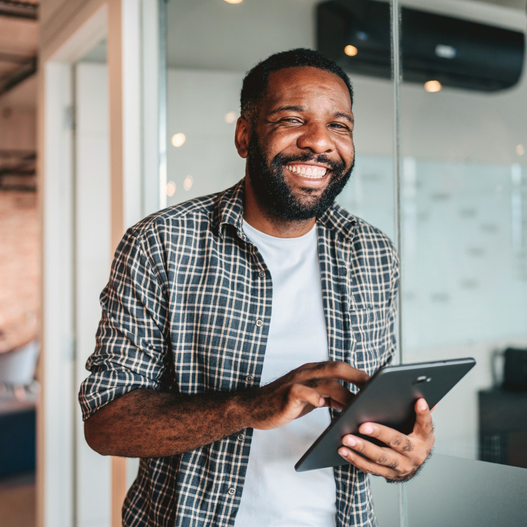 A smiling man in a plaid shirt using a tablet in a modern office setting.