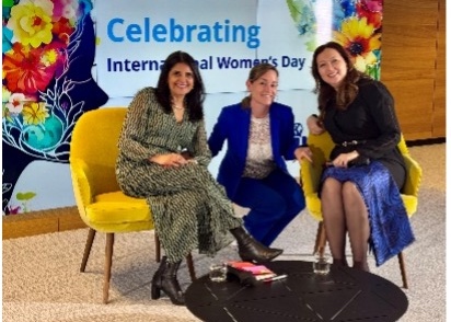 Three women sitting and posing for photo in front of Celebrating International Women's Day banner