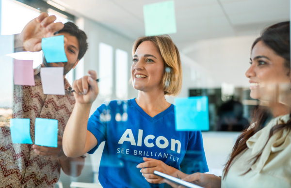 Three colleagues collaborating at a glass wall covered in colorful sticky notes; the woman in the center wears an Alcon branded shirt.