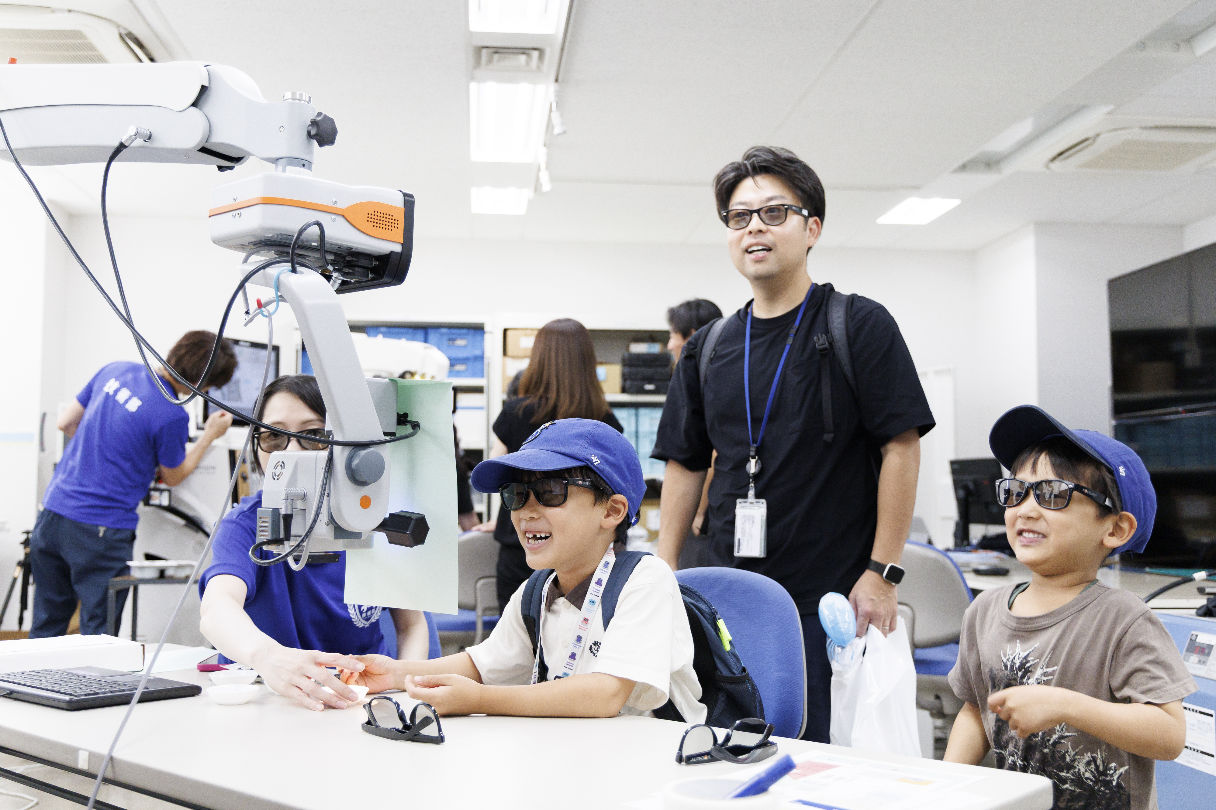 明るいクリニックで、二人の子どもがスタッフと笑顔の男性に案内されながら、視力検査機器を興味深そうに見ている