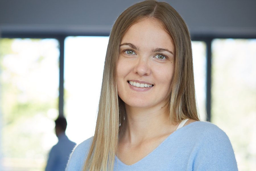 Eine Frau mit langen Haaren in einem blauen Pullover lächelt freundlich in einem sonnendurchfluteten Büro.