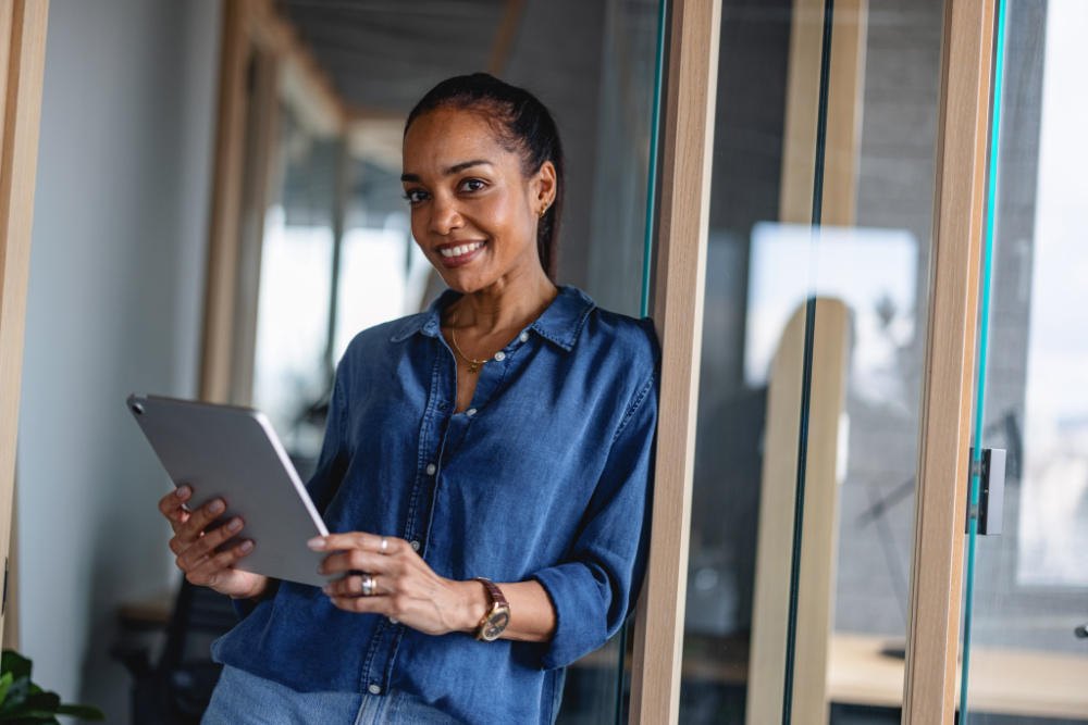A smiling woman is looking down at a tablet she is holding in both hands. She is wearing a blue button-down shirt and a watch.
