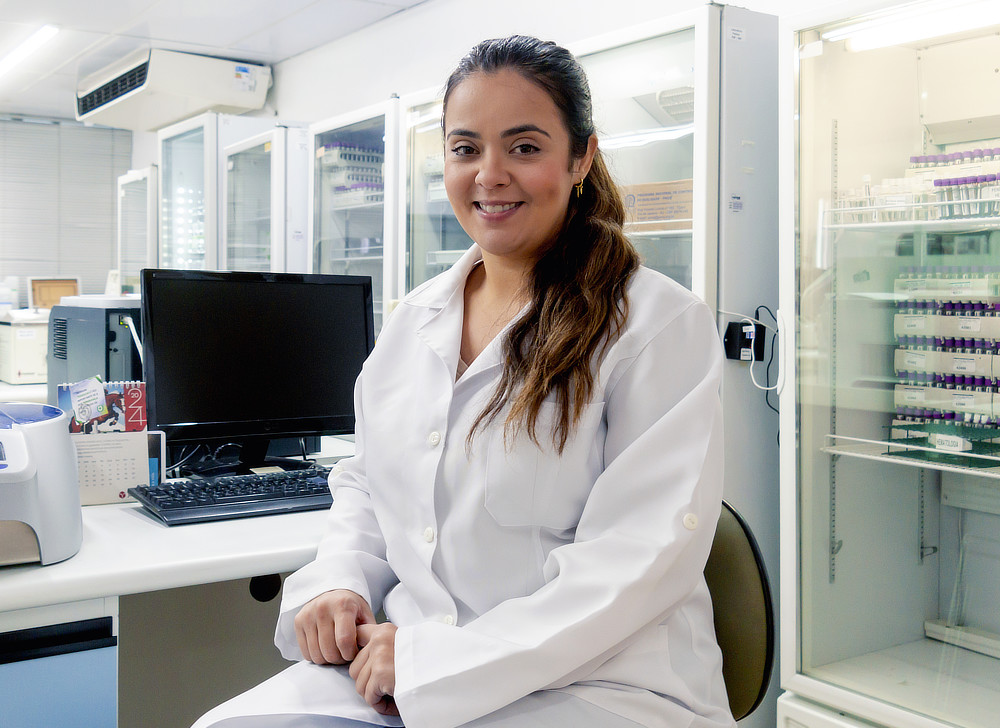 A woman with long brown hair is smiling at the camera while seated at a lab bench. She is wearing a white lab coat and is surrounded by lab equipment.