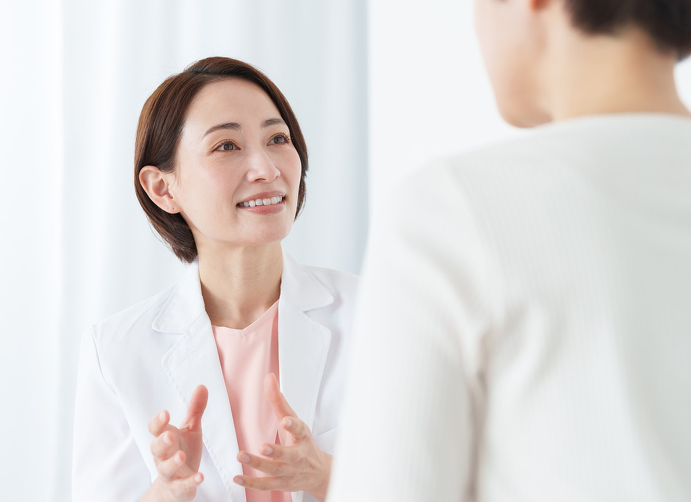 A female doctor wearing a white coat is smiling and talking to a patient. The patient is partially visible, facing away from the camera.