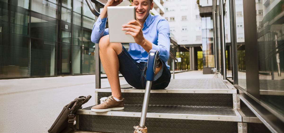 Young man with a prosthetic leg, sitting on stairs outside office building, smiling and looking at tablet