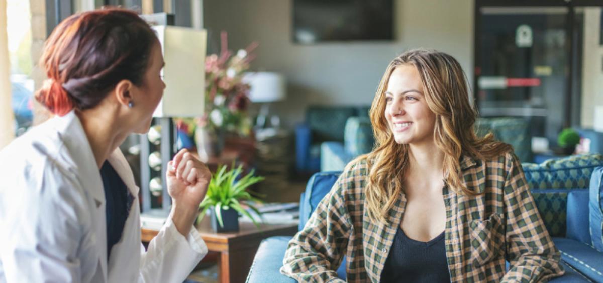 A healthcare professional in a white coat engages in conversation with a smiling woman in a plaid shirt, sitting in a cozy, well-lit waiting area with green plants and comfortable furniture.