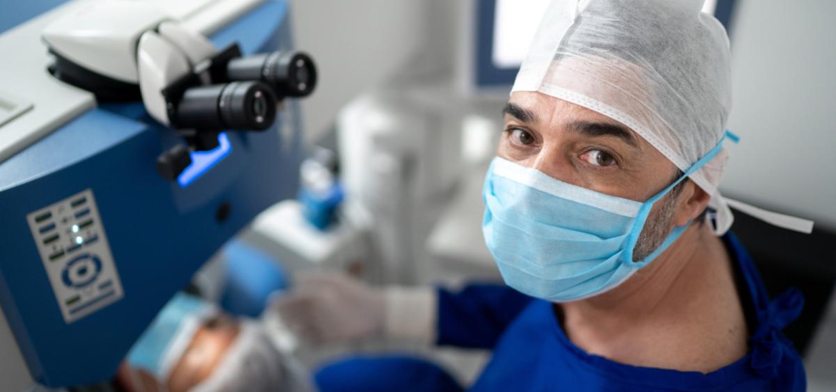 A close-up of an eye surgeon wearing a surgical cap and mask, looking directly at the camera with an eye scope in the background.