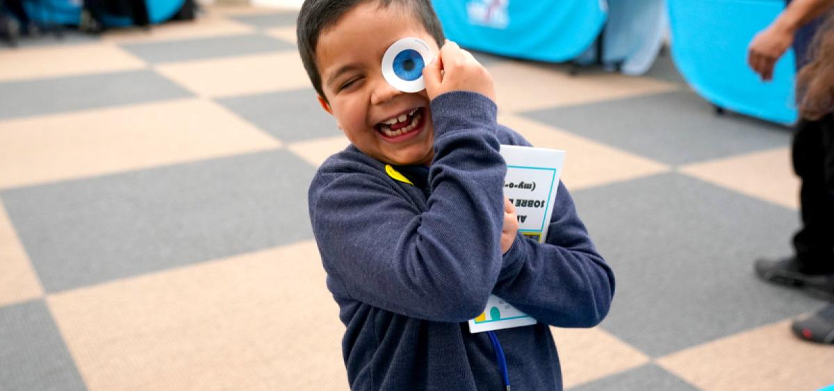 Smiling child holding a large googly eye toy up to their face while clutching a book outdoors.