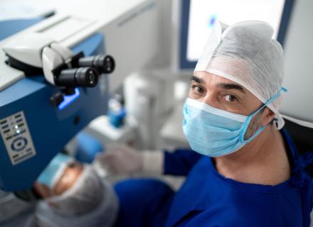 A close-up of an eye surgeon wearing a surgical cap and mask, looking directly at the camera with an eye scope in the background.