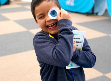 Smiling child holding a large googly eye toy up to their face while clutching a book outdoors.