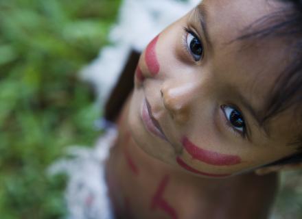 A child's face with red painted stripes on the cheeks is looking upward.