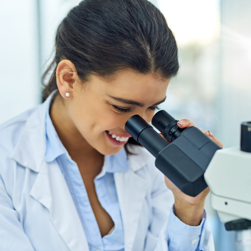 Woman in lab coat looking through microscope and smiling.
