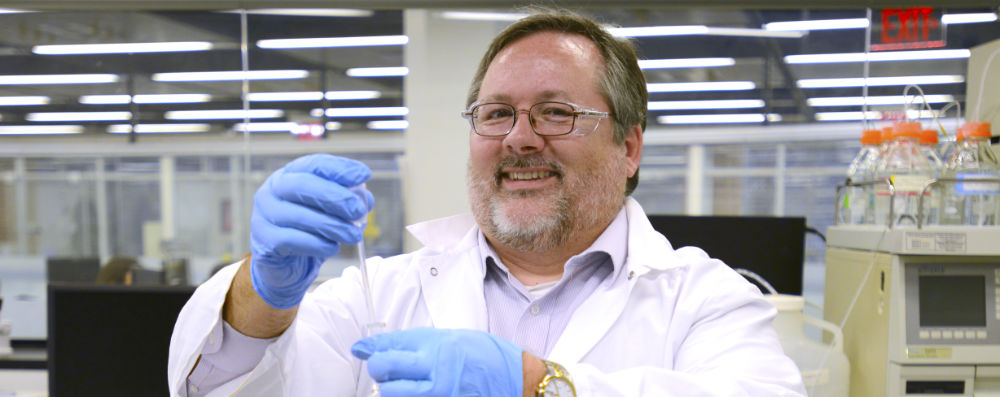 A smiling man wearing blue nitrile gloves, and wearing a white lab coat, holding a pipette.
